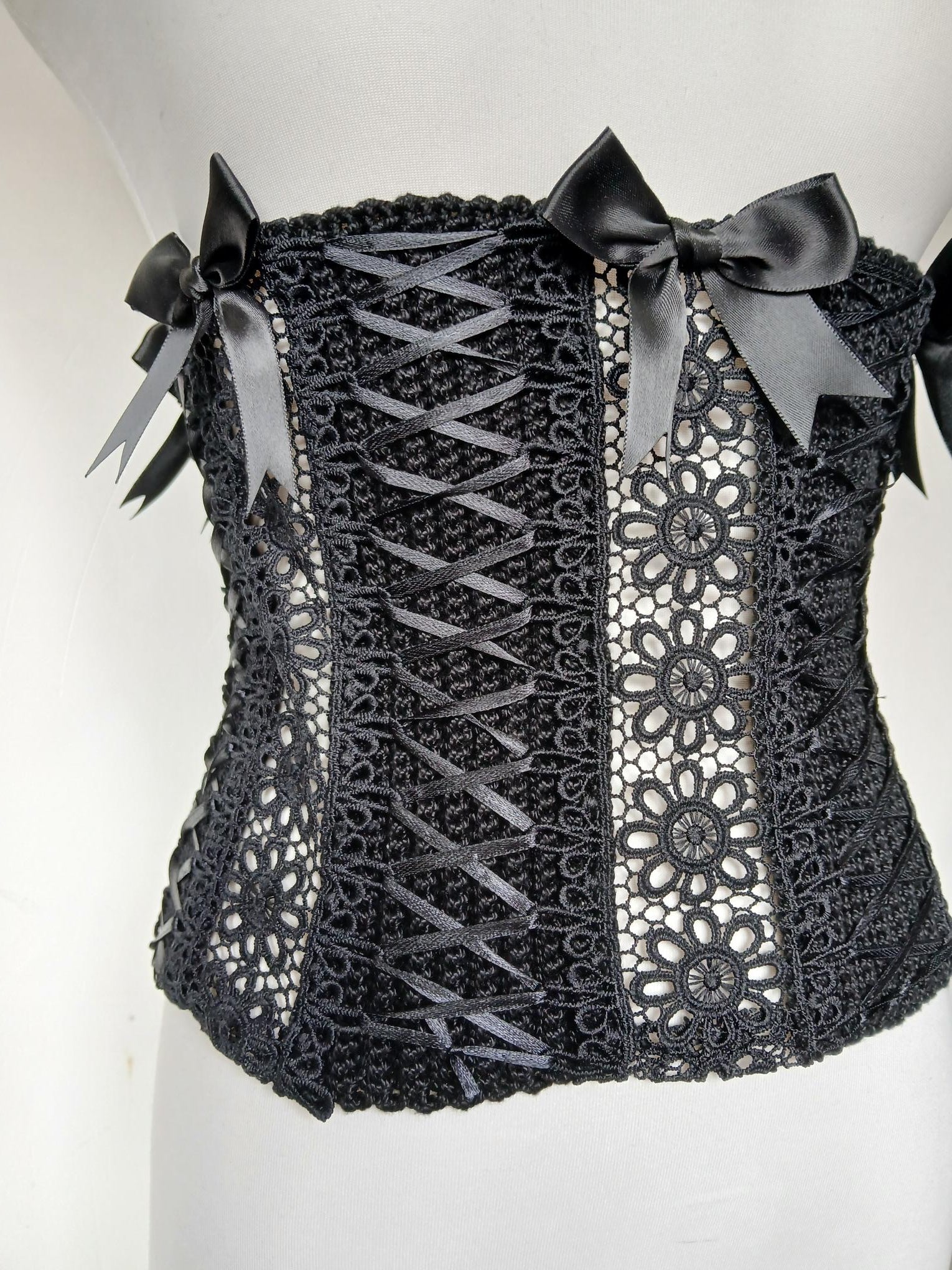 Gothic Lace Corset Belt The Dark Duchess — close up detail of black handmade crochet corset with guipure lace panel, satin ribbon criss-cross lacing and black satin bows on white mannequin - Victorian Velvet