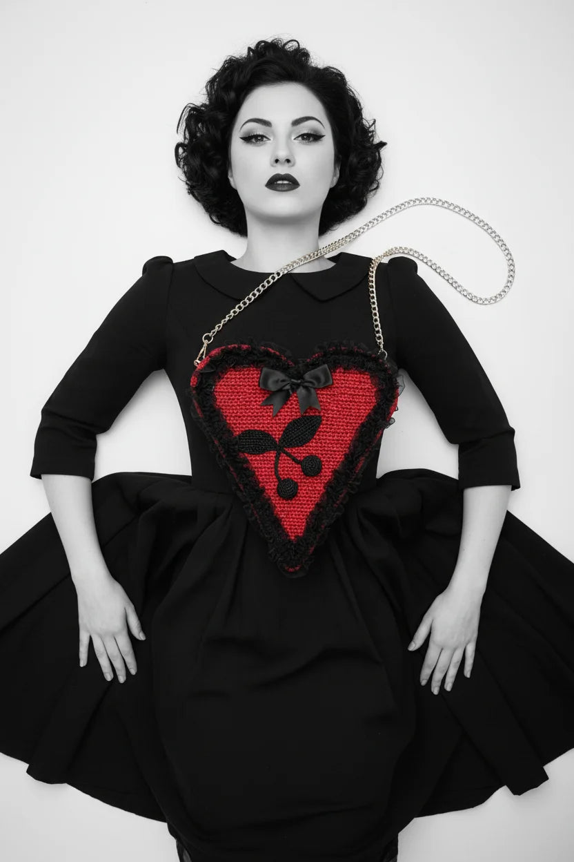 Overhead view of pin-up model in black dress lying down, red crochet heart bag on chest, black lace trim, satin bow and cherry appliqué, gold chain strap beside her — Victorian Velvet