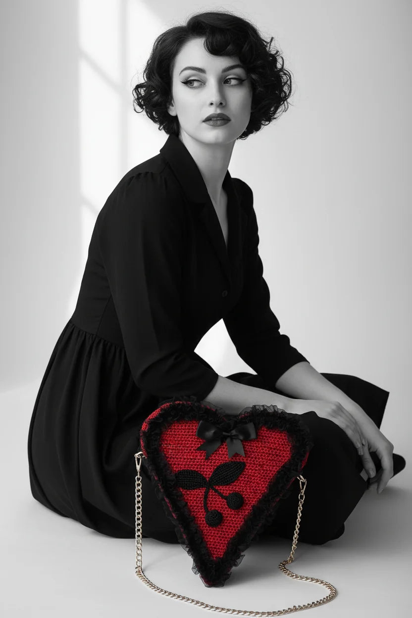 Pin-up model in black dress sitting on floor, red crochet heart-shaped bag resting on lap, black lace trim, satin bow and cherry appliqué detail, gold chain strap — Victorian Velvet