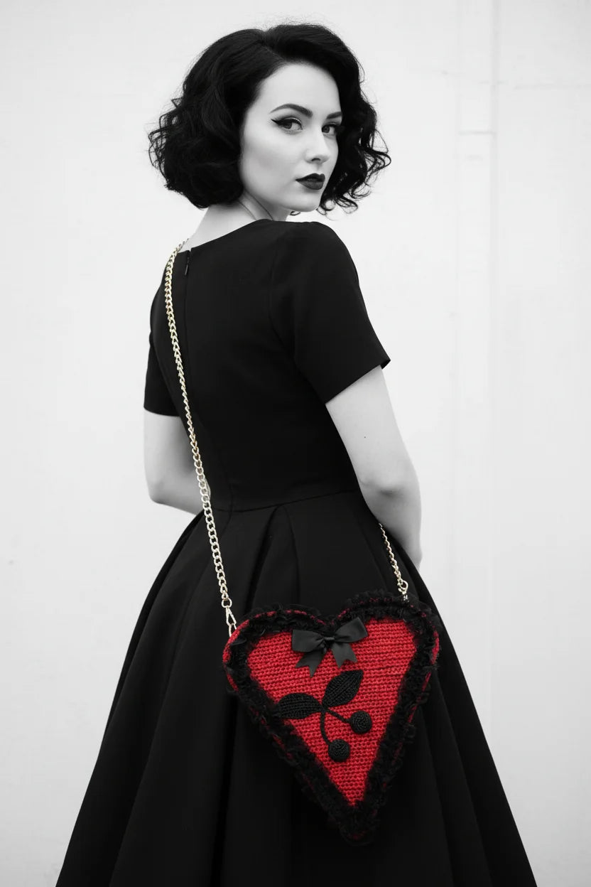 Pin-up model in black dress photographed from behind, looking over shoulder, red crochet heart bag with cherry appliqué and black lace trim worn on gold chain strap — Victorian Velvet