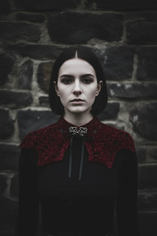 Pale gothic woman with short dark hair wearing a deep burgundy velour crochet bat wing collar with silver baroque brooch and black satin ribbon, standing against dark stone ruins, gothic fashion editorial - Victorian Velvet