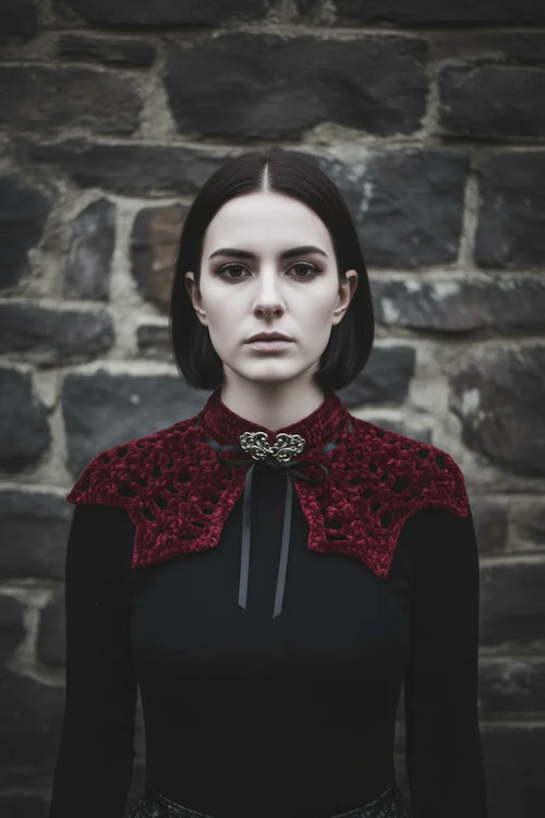 Pale gothic woman with short dark bob hair wearing a deep burgundy velour crochet bat wing collar with silver baroque brooch and black satin ribbon, standing against grey stone wall, gothic fashion editorial - Victorian Velvet