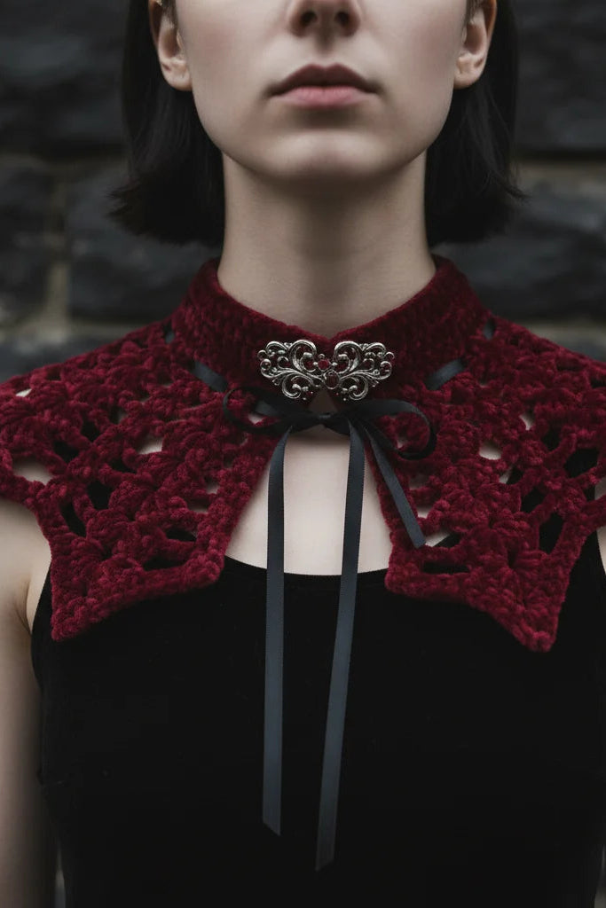 Close-up of a deep burgundy velour crochet bat wing collar with open lace pattern, silver baroque brooch and black satin ribbon tie at center front, worn against dark stone background - Victorian Velvet