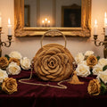 Handmade golden crochet rose handbag with gold ring handle, gold chain strap and golden tassel, displayed on dark burgundy velvet surrounded by white and golden roses, antique gold mirror and candelabras in the background by Victorian Velvet