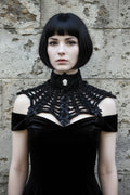 Front view of a pale gothic woman with short dark black bob hair with fringe wearing a black chunky velour chenille crochet collar capelet with high turtleneck band, large oval black cameo brooch with white Victorian lady silhouette profile at center front, wide bat wing shaped open spider web mesh crochet panel with small black satin ribbon bows at yarn intersections, worn over a black off-shoulder velvet dress, grey stone wall background - Victorian Velvet