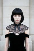 Pale gothic woman with short dark bob hair wearing a black sheer tulle lace collar capelet with embroidered gothic cross motifs, scalloped lace edges and black crystal brooch at center front, worn over a black off-shoulder velvet dress against grey stone wall - Victorian Velvet