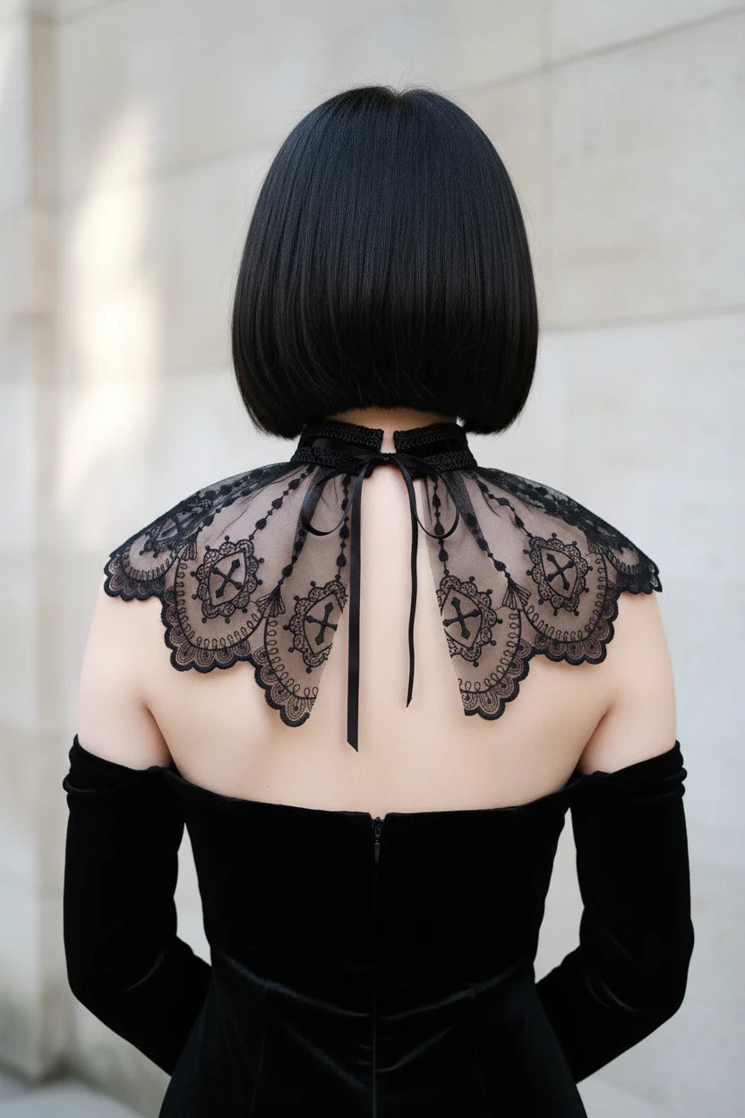 Back view of a pale gothic woman with short dark bob hair wearing a black sheer tulle lace collar capelet with embroidered gothic cross motifs and scalloped edges, black velvet choker band with ribbon tie at center back, worn over a black off-shoulder velvet dress - Victorian Velvet
