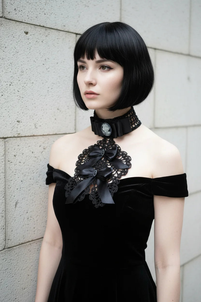 Pale gothic woman with short dark bob hair at a three-quarter angle wearing a black Victorian jabot with grey cameo brooch and cascading black crochet lace bib with black satin bows, worn over a black off-shoulder velvet dress, grey stone wall background - Victorian Velvet