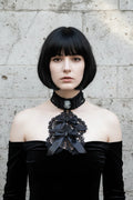 Pale gothic woman with short dark bob hair wearing a black Victorian jabot with grey cameo brooch, black crochet lace bib and black satin bows with crystal brooches, worn over a black off-shoulder velvet dress with bare shoulders, grey stone wall background - Victorian Velvet