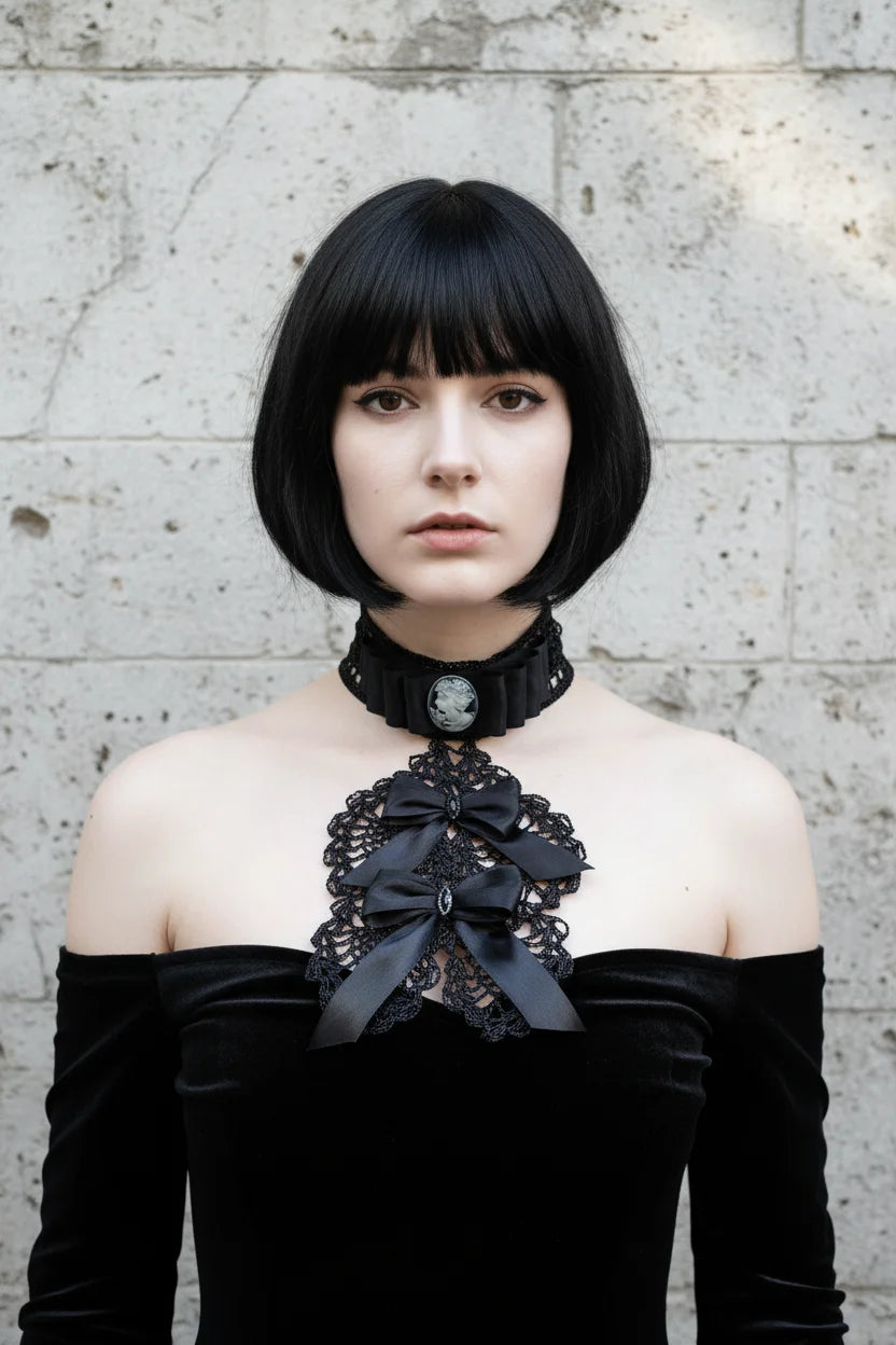 Pale gothic woman with short dark bob hair wearing a black Victorian jabot with grey cameo brooch, black crochet lace bib and black satin bows with crystal brooches, worn over a black off-shoulder velvet dress with bare shoulders, grey stone wall background - Victorian Velvet