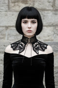 Front view of a pale gothic woman with short dark black bob hair with fringe wearing a black fine crochet lace collar with wide high turtleneck band with open square grid mesh pattern and black corset-style lace-up ribbon tie at center front, wide dramatic wing-shaped lace crochet panels draping over both shoulders with intricate gothic lace pattern and scalloped pointed tips, worn over a black off-shoulder velvet dress, grey stone wall background - Victorian Velvet