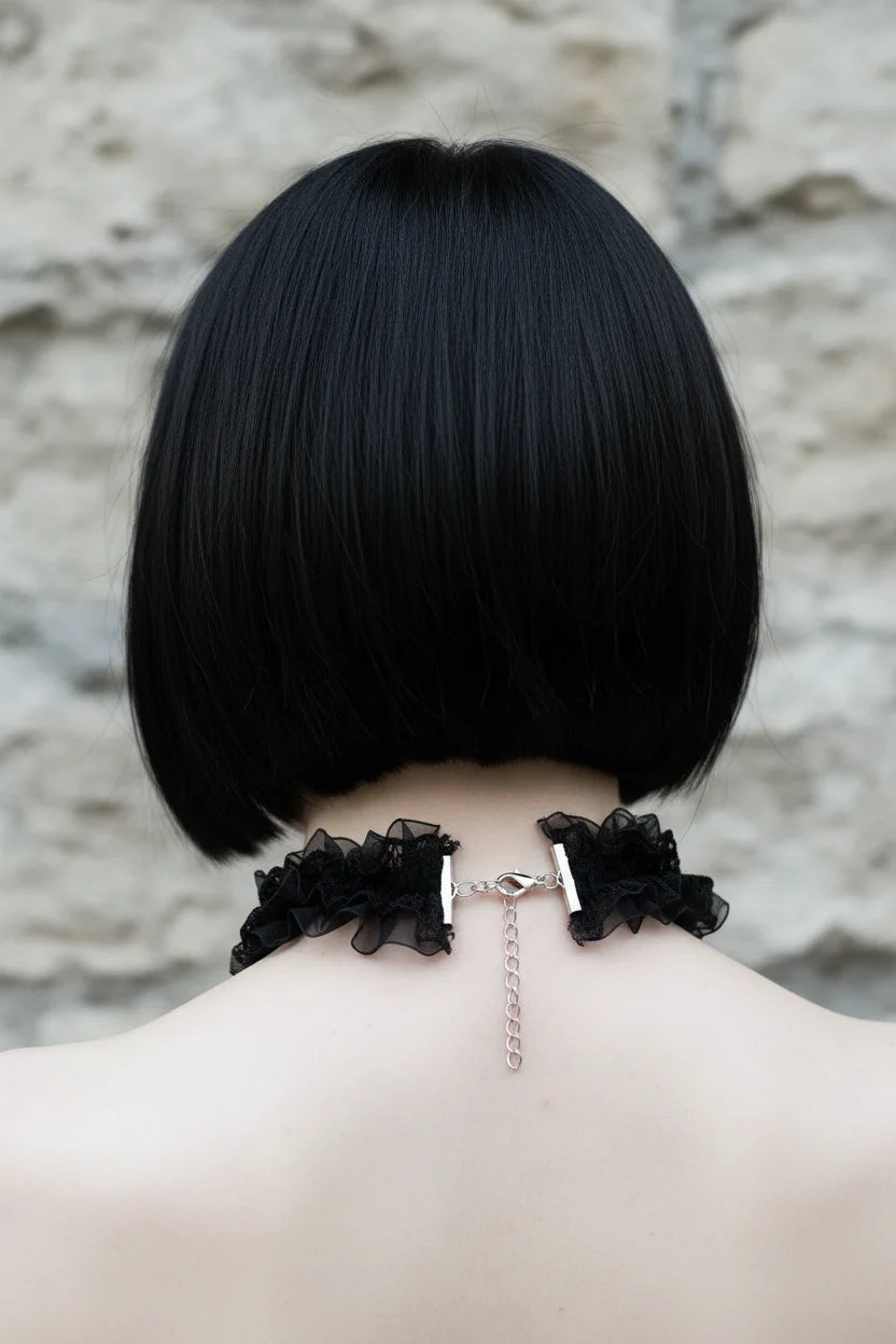 Back view close-up of a pale gothic woman with short dark black bob hair with fringe wearing a black gothic choker necklace made of layered black organza and black lace ruffles with silver lobster clasp and short silver extension chain closure at center back, grey stone wall background - Victorian Velvet