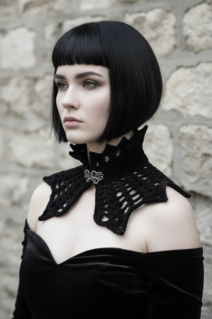 Side view of a pale gothic woman with short dark black bob hair with fringe wearing a short black velour crochet collar capelet with pointed crown spikes at the turtleneck, ornate antique silver baroque filigree brooch clasp visible at center front, and wide bat wing shaped crochet panel with open diagonal mesh pattern and pointed tips, worn over a black off-shoulder velvet dress, grey stone wall background - Victorian Velvet