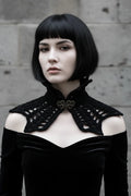 Pale gothic woman with short dark black bob hair with fringe wearing a short black velour crochet collar capelet with pointed crown spikes at the turtleneck and ornate antique bronze baroque filigree brooch clasp at center front, wide bat wing crochet panels with open diagonal mesh pattern, worn over a black off-shoulder velvet dress, grey stone wall background - Victorian Velvet