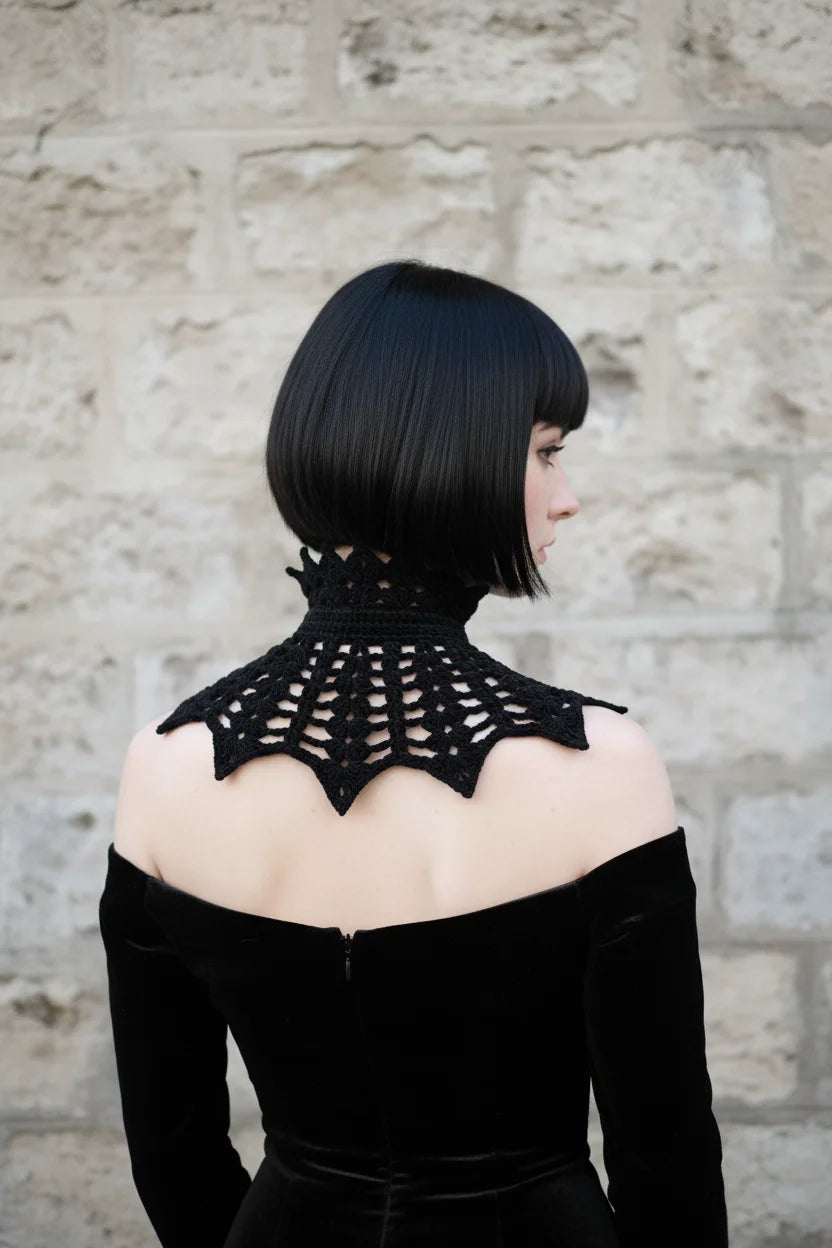 Back view of a pale gothic woman with short dark black bob hair with fringe wearing a short black velour crochet collar capelet with pointed crown spikes at the turtleneck and wide symmetrical bat wing crochet panels with open spider web mesh pattern and pointed tips at the bottom edges, worn over a black off-shoulder velvet dress, stone wall background - Victorian Velvet