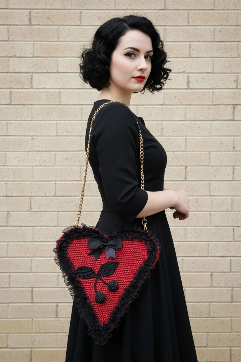 Pin-up model in black dress wearing the Poison Cherry Heart Bag on a gold chain strap, side profile view against a light brick wall - Victorian Velvet