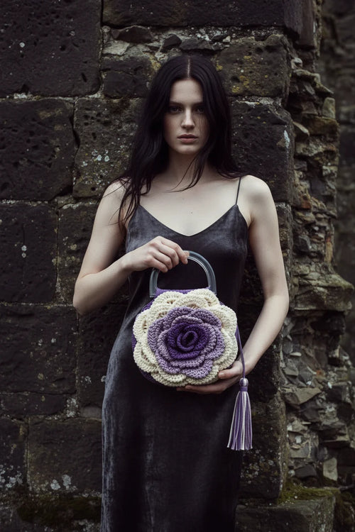 Woman with dark hair in a dark velvet slip dress holding a cream and purple crochet rose handbag with clear acrylic handle and lavender tassel, standing against a rough dark stone wall, gothic fashion editorial - Victorian Velvet