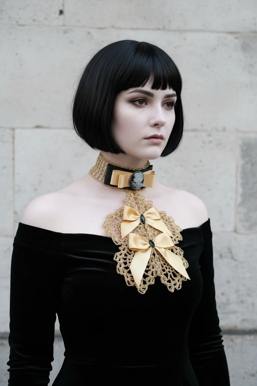 Pale gothic woman with short dark black bob hair with fringe at three-quarter angle wearing a gold metallic crochet Victorian jabot with black velvet choker band, grey cameo brooch, gold satin bow and cascading gold lace bib with gold satin bows and black crystal brooches, worn over a black off-shoulder velvet dress, grey stone wall background - Victorian Velvet