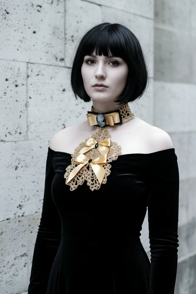 Pale gothic woman with short dark black bob hair with fringe wearing a gold metallic crochet Victorian jabot with black velvet choker band, grey cameo brooch, gold satin bow and cascading gold lace bib with two gold satin bows and black crystal brooches, worn over a black off-shoulder velvet dress, grey stone wall background - Victorian Velvet