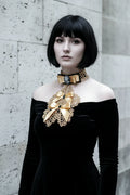 Pale gothic woman with short dark black bob hair with fringe wearing a gold metallic crochet Victorian jabot with black velvet choker band, grey cameo brooch, gold satin bow and cascading gold lace bib with two gold satin bows and black crystal brooches, worn over a black off-shoulder velvet dress, grey stone wall background - Victorian Velvet
