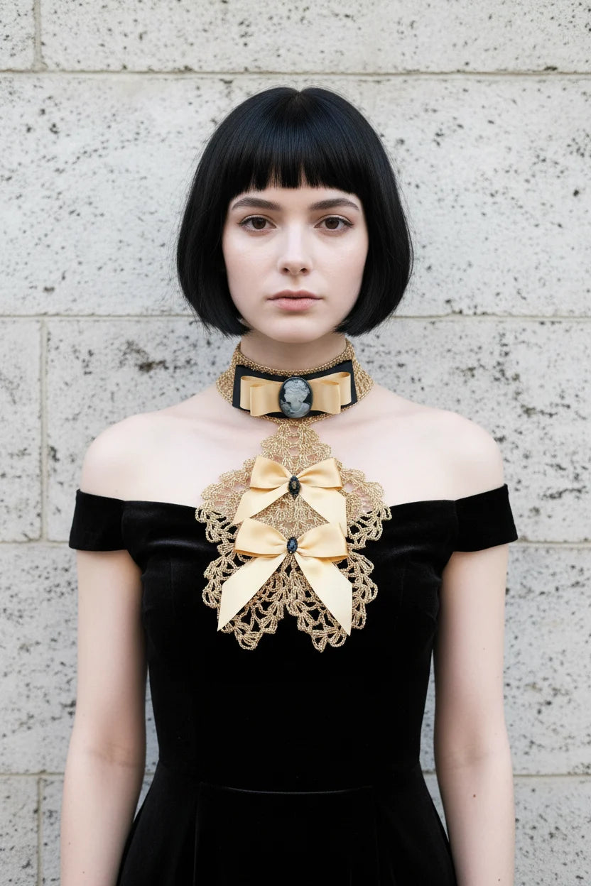 Pale gothic woman with short dark black bob hair with fringe wearing a gold metallic crochet Victorian jabot with black velvet choker band, grey cameo brooch, gold satin bow and cascading gold lace bib with two gold satin bows and black crystal brooches, worn over a black off-shoulder velvet dress, grey stone wall background - Victorian Velvet