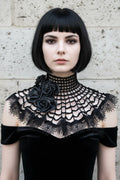 Front view of a pale gothic woman with short dark black bob hair with straight fringe wearing a black crochet spider web collar capelet over a black off-shoulder velvet dress, high fitted open grid mesh turtleneck band, wide circular capelet with spider web crochet pattern, cluster of three large black satin roses with small silver spider brooches on the left side, wide black lace ruffle trim with bead drops at the outer edge, grey stone wall background - Victorian Velvet
