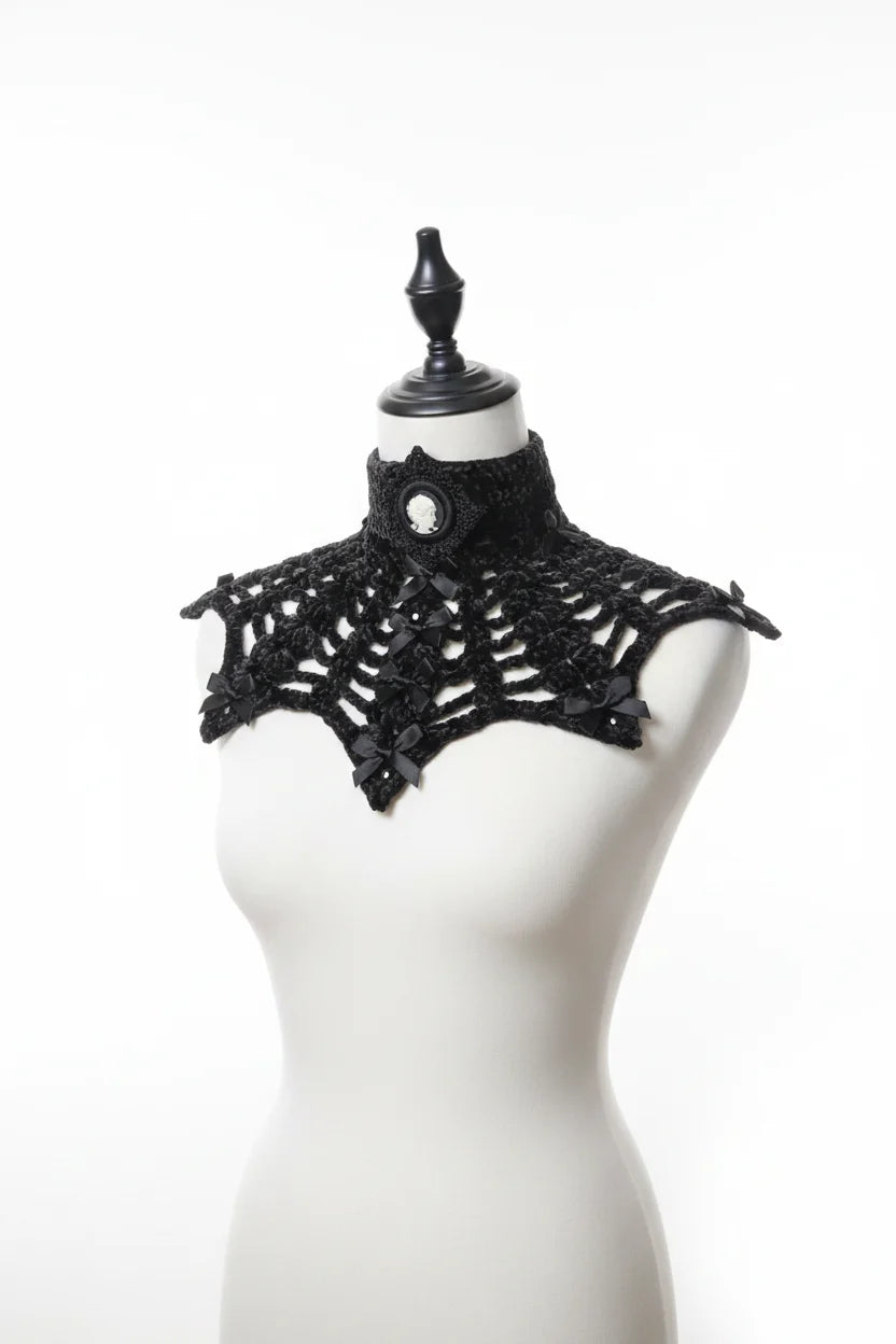 Front view of a black chunky velour chenille crochet collar capelet on a white dress form mannequin, high turtleneck band with large oval black cameo brooch with white Victorian lady silhouette profile at center front, wide bat wing shaped open spider web mesh crochet panel with small black satin ribbon bows at yarn intersections and pointed tips at outer shoulder edges and bottom center, pure white background - Victorian Velvet