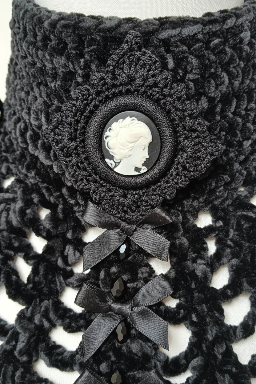 Close-up detail of a black chunky velour chenille crochet collar showing the large oval black ornate crocheted frame with cameo brooch depicting a white Victorian lady silhouette profile facing left with curly updo hair, below the cameo open spider web mesh crochet panel with small black satin ribbon bows and black crystal beads at yarn intersections, white background - Victorian Velvet