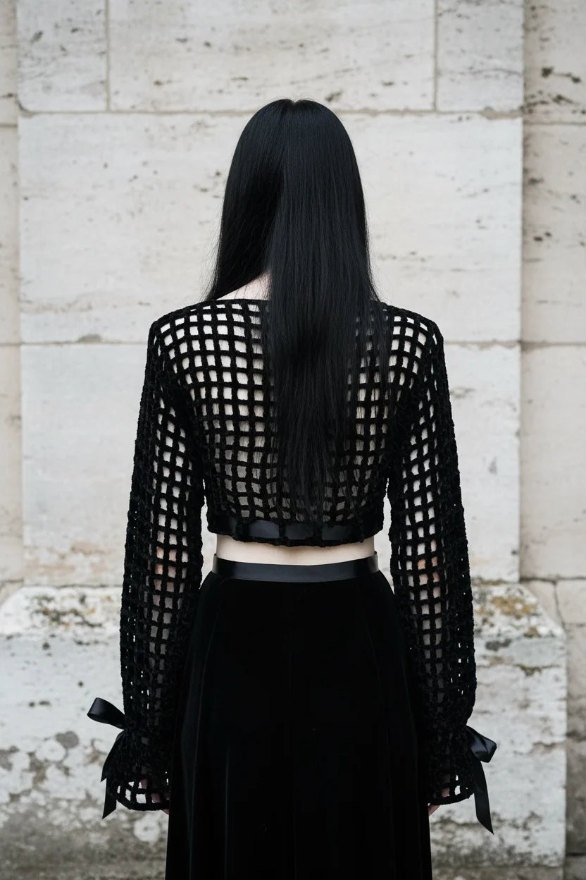 Black velour grid crochet crop top back view showing open grid pattern and long sleeves with satin bow ties at wrists, gothic model with long dark hair, stone wall background - Victorian Velvet